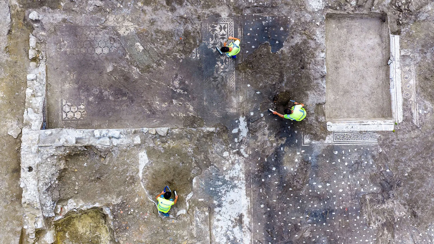 an aerial photograph of three archaeologists in high-viz vests excavating in four rooms of an ancient villa with mosaics on the floor