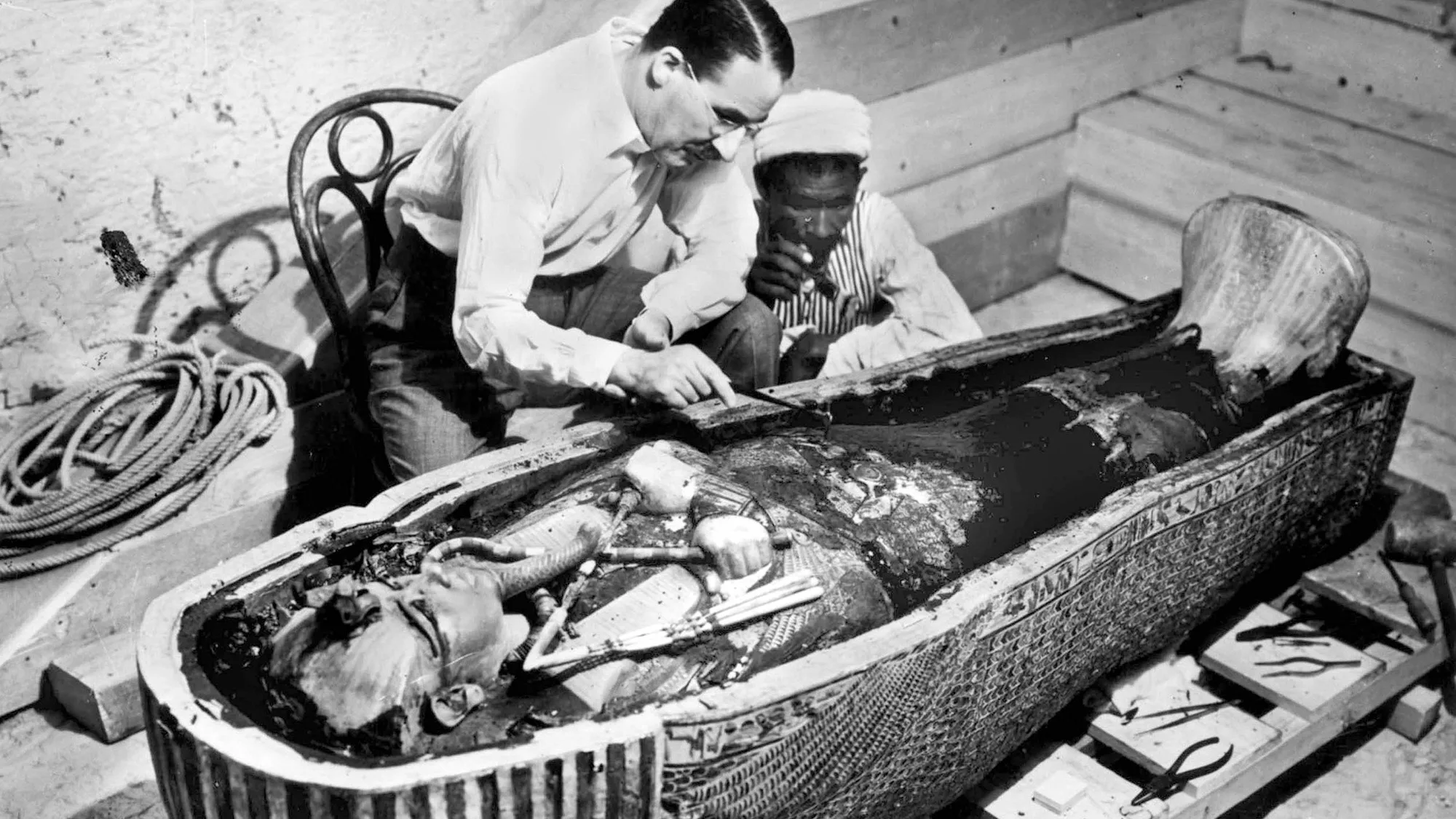 A black and white photo of two men peering over an open sarcophagus.
